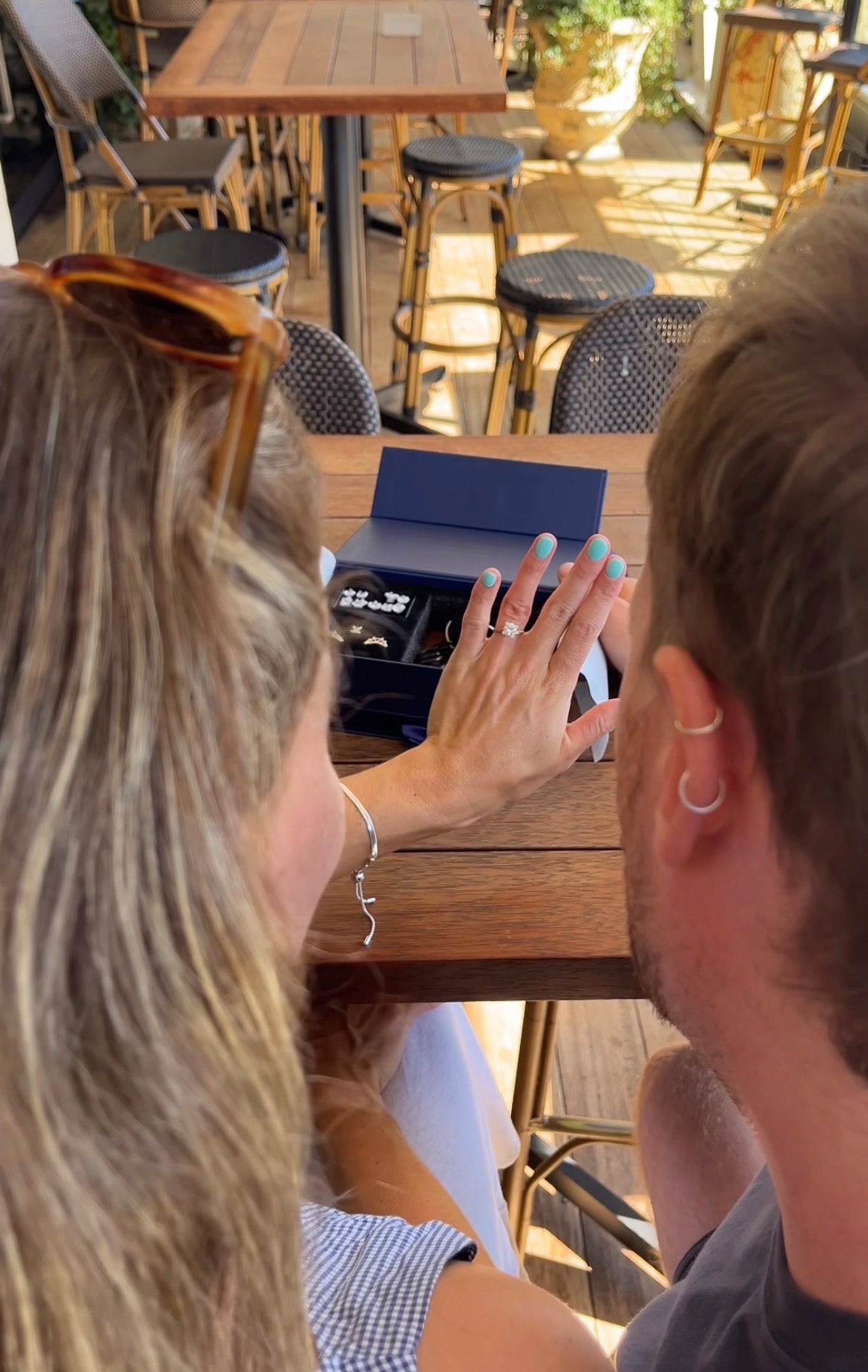 Woman's left hand displaying engagement ring with teal nails over open jewelry box on a cafe table, man beside her