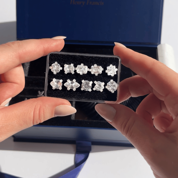 Hands holding clear tray of ten diamond-cut stud earrings in varied shapes inside an open blue Henry Francis jewelry box