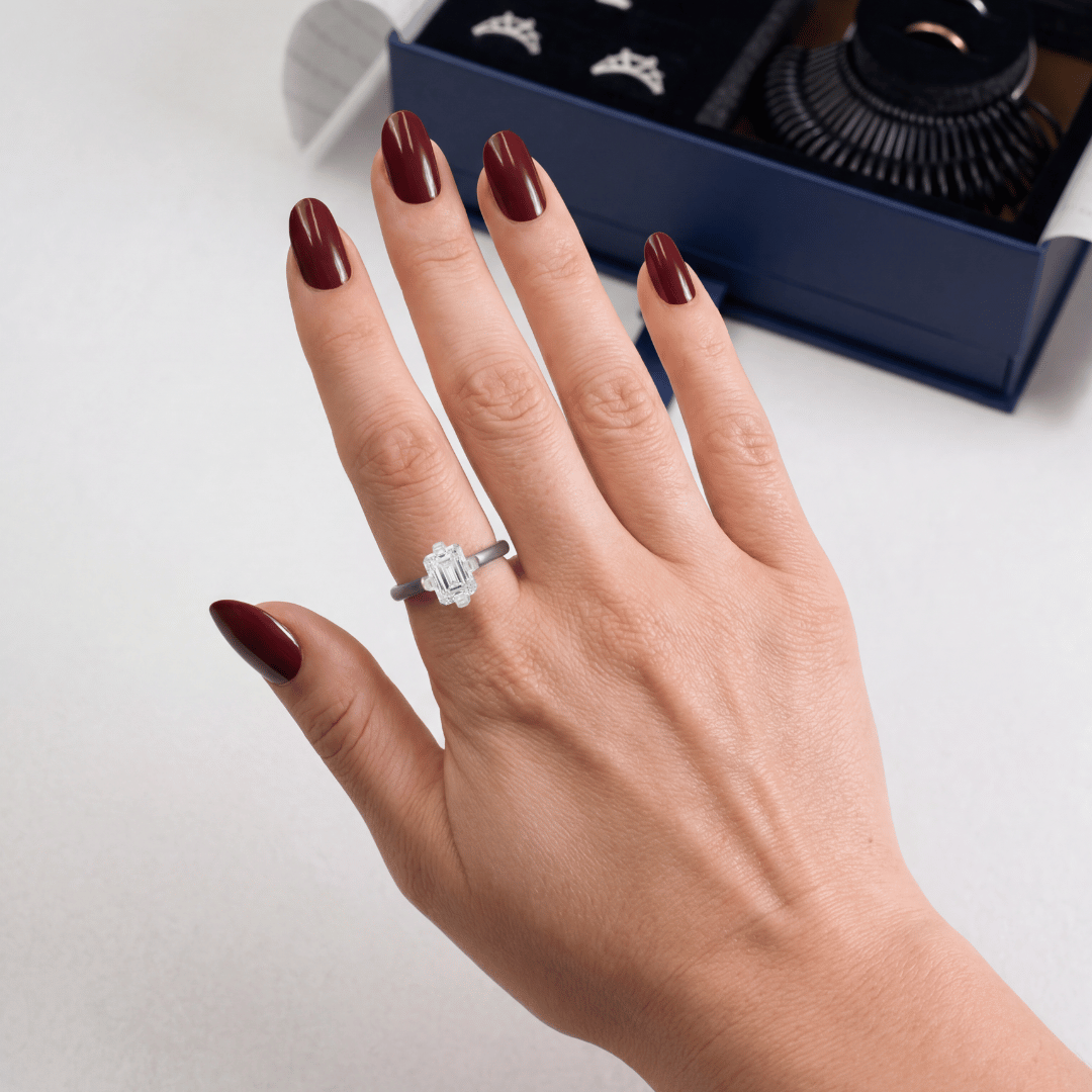 Close-up of hand wearing emerald-cut engagement ring showing emerald-cut stone and prong setting with navy try-on kit box