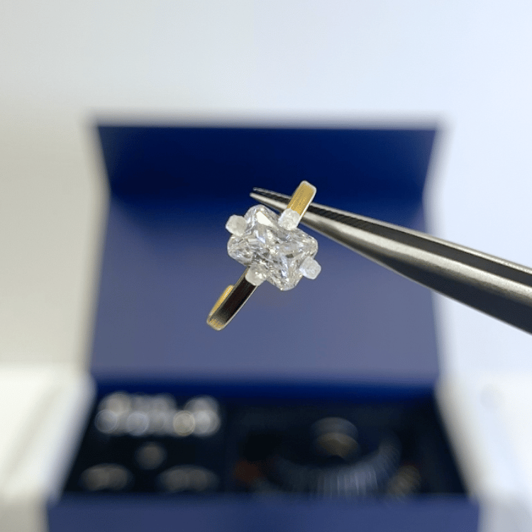 Close-up of a replica clear stone on a gold-tone try-on band held with tweezers above an open blue Henry Francis kit box