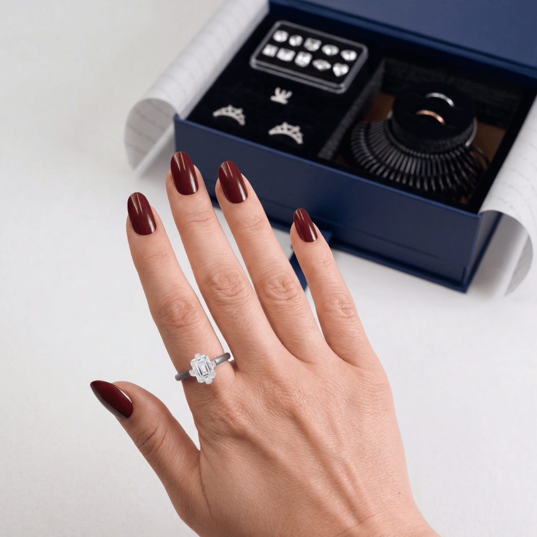 Hand wearing an emerald-cut engagement ring with dark red nails in front of an open navy Ring Try-On Kit box showing ring shapes and bands