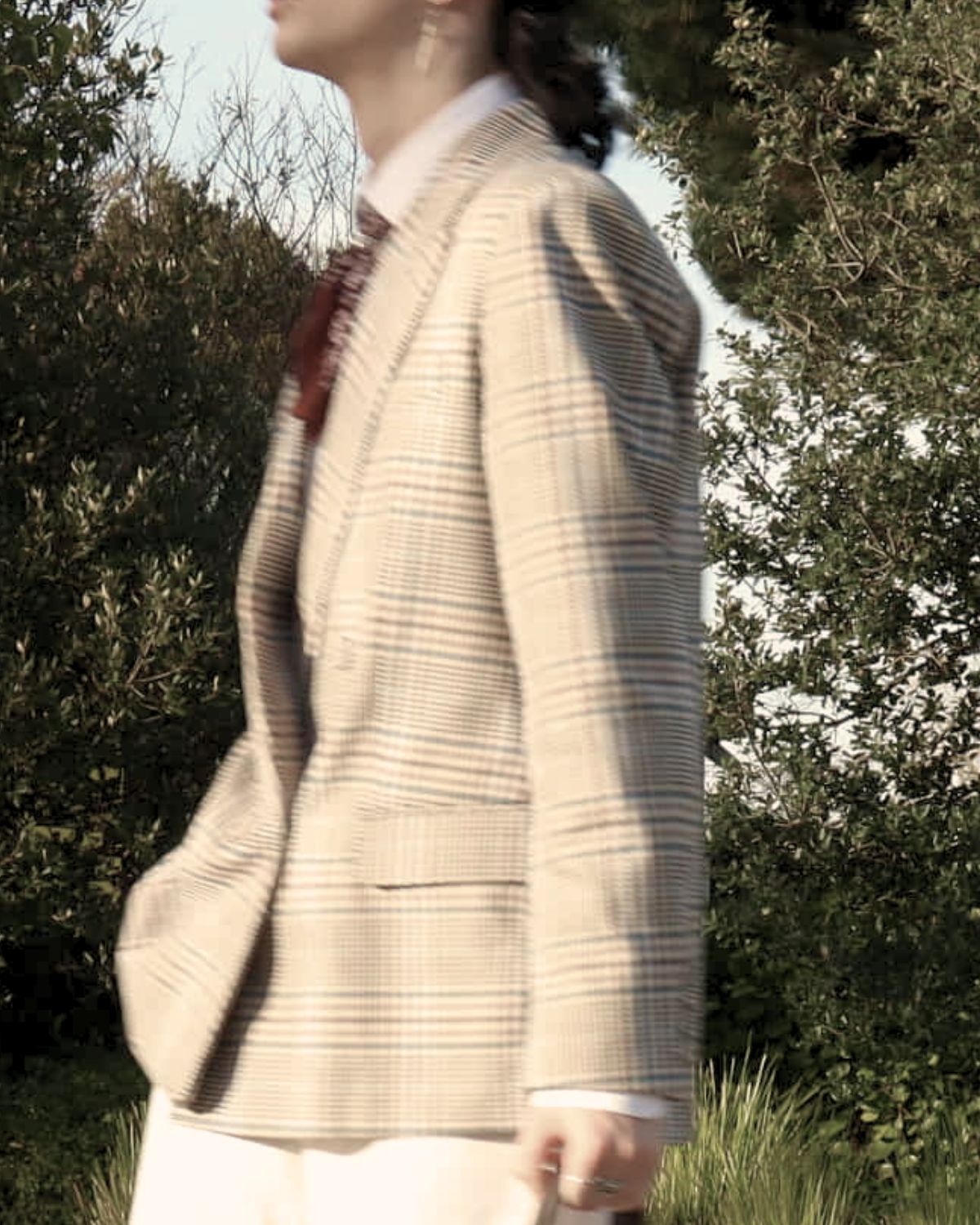 Model in a check blazer and white trousers walking through a garden, wearing a gold drop earring