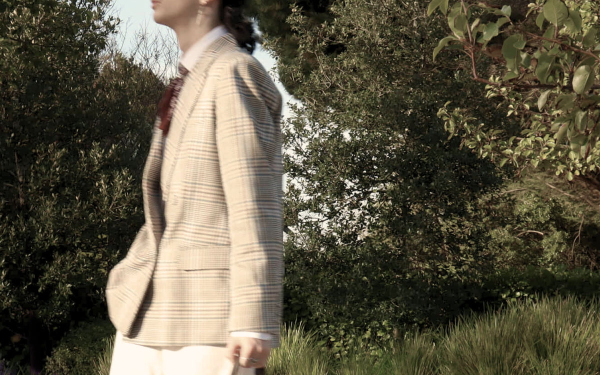 Model in a check blazer walking past native bush in soft daylight