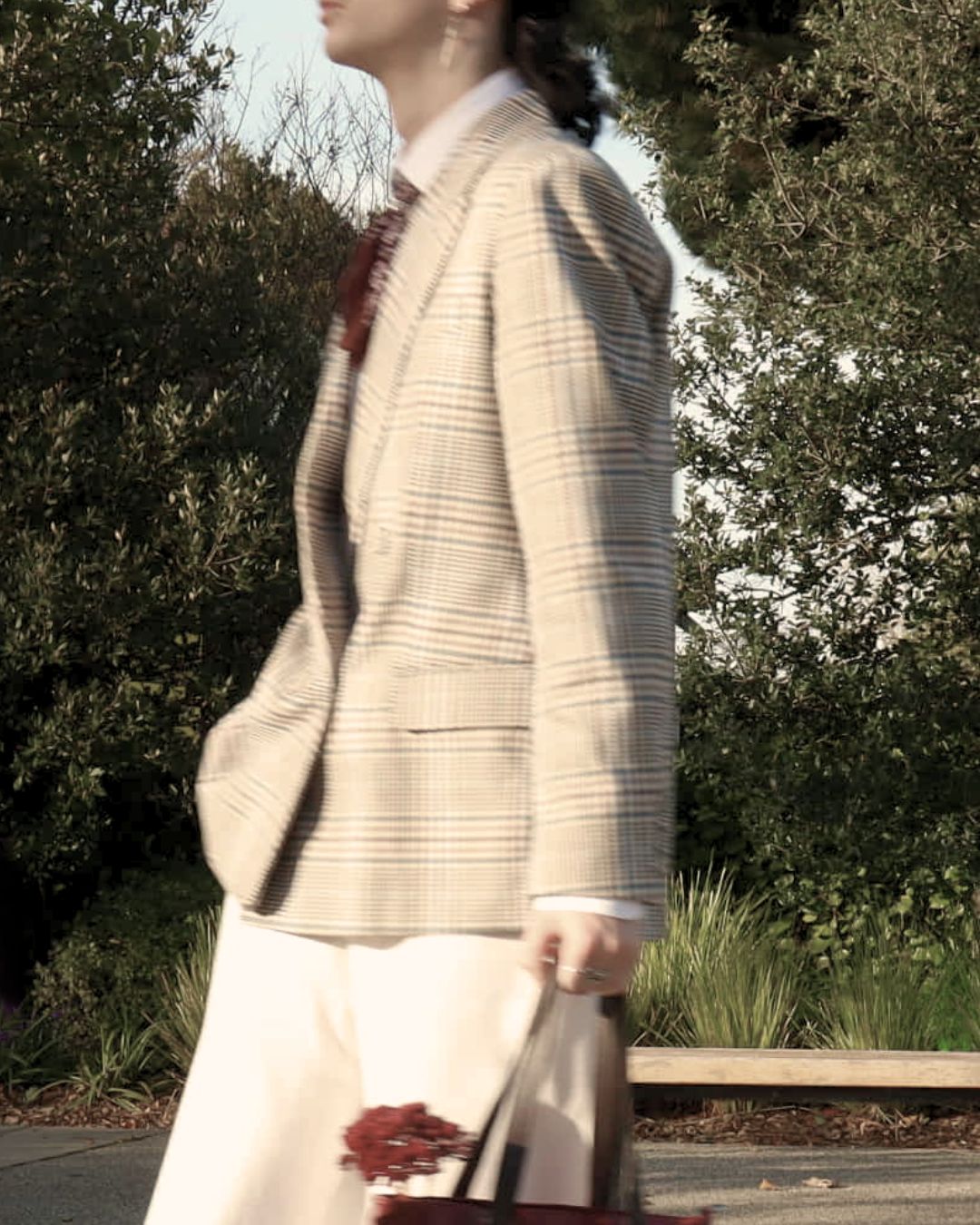 Model in a check blazer carrying a bag of red flowers through a garden